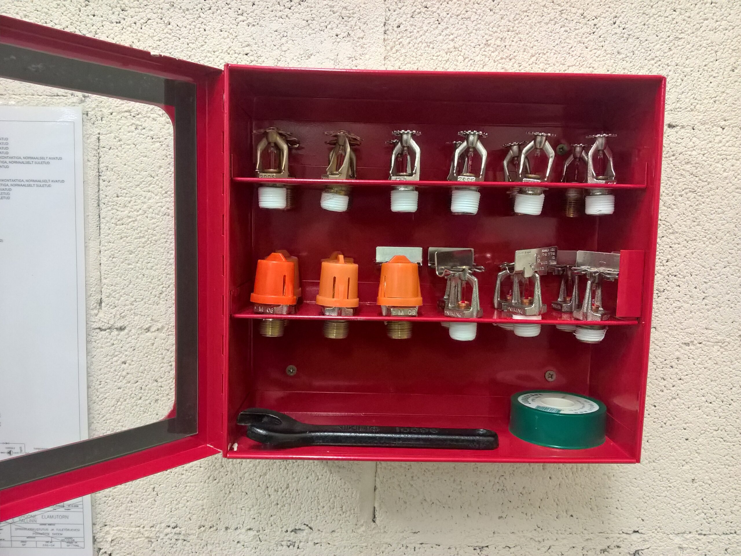 Avatud punane metallist sprinkleri varuosade kapp, milles on kaks riiulit erinevate sprinklerpeadega, paigaldusvõti ja keermeteip, kinnitatud betoonplokk-seinale.